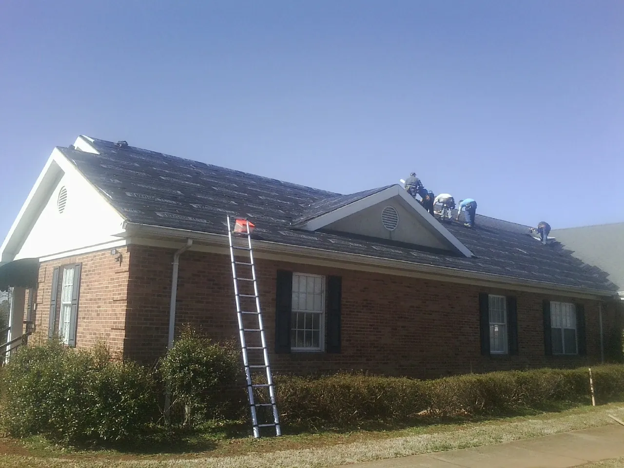 Crew tearing shingles off a brick ranch roof with a ladder set — Metal Roof Repair work by our crew in Emery